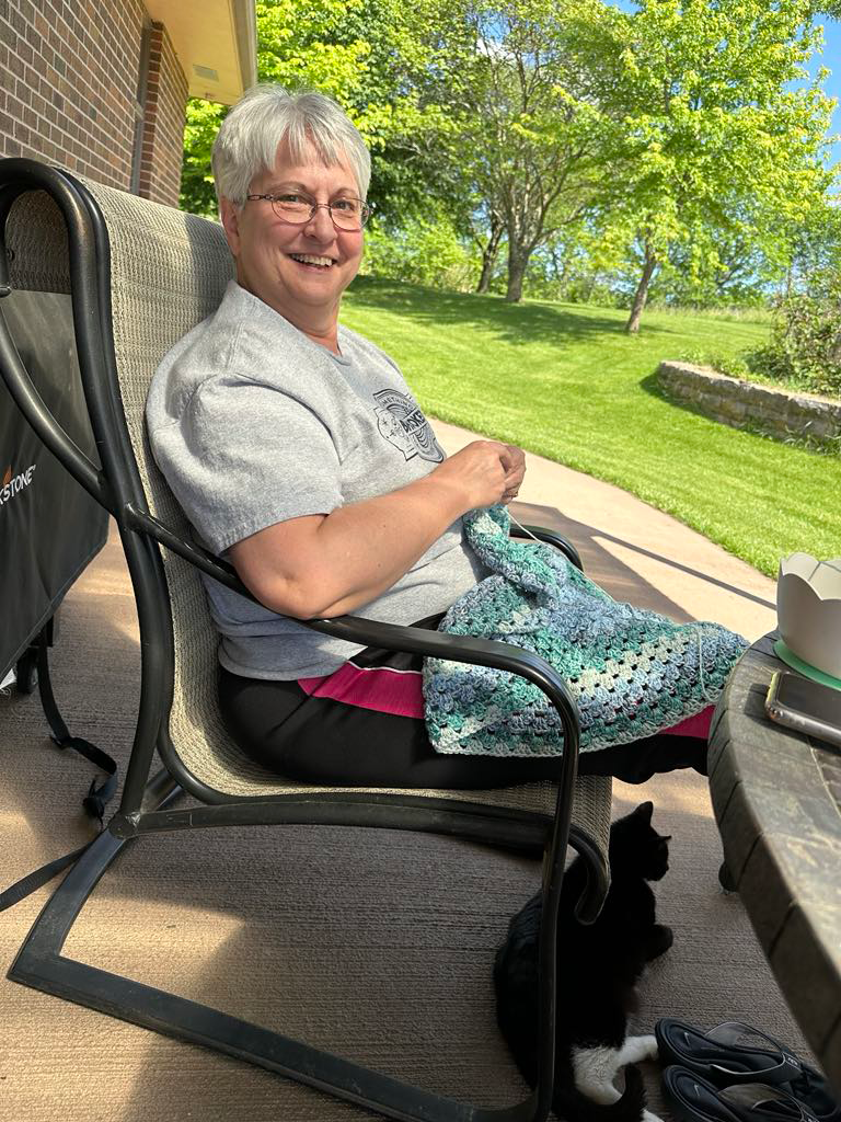 Belle crocheting outside while her cat Bandit sits under her chair